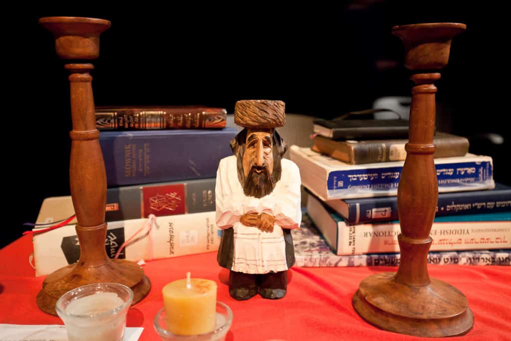 A picture of a small wooden Jew in between two wooden Shabbat candlesticks. Behind them, there is a stack of books.